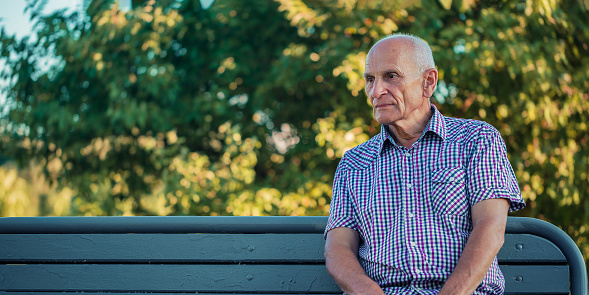 Solitary sad senior man with gray hair sitting on wooden bench against autumn leaves and thinking