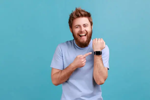 Portrait of positive funny bearded man standing with open mouth and satisfied expression, pointing with finger ar smartwatch and laughing. Indoor studio shot isolated on blue background.