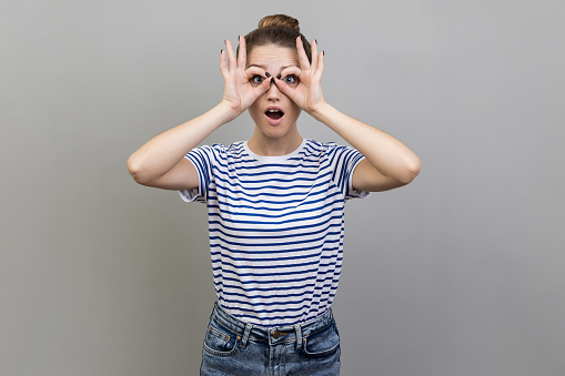 Portrait of woman wearing striped T-shirt looking through binoculars gesture and expressing surprise, zooming vision, exploring distance. Indoor studio shot isolated on gray background.