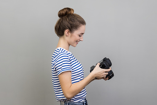 Side view portrait of satisfied delighted woman traveler or photographer wearing striped T-shirt holding and looking at photocamera, taking photo enjoying hobby. Indoor studio shot isolated on gray background.