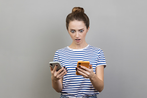 Portrait of shocked astonished woman wearing striped T-shirt using two smart phones, looking at display with big eyes, being surprised. Indoor studio shot isolated on gray background.