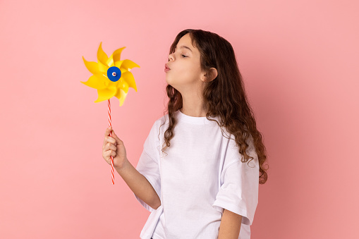Portrait of charming cute playful little girl wearing white T-shirt blowing at paper windmill, playing with pinwheel toy on stick. Indoor studio shot isolated on pink background.