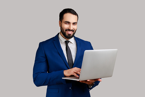 Positive bearded man holding laptop, looking at camera, surfing internet, freelancer working using computer, wearing official style suit. Indoor studio shot isolated on gray background.