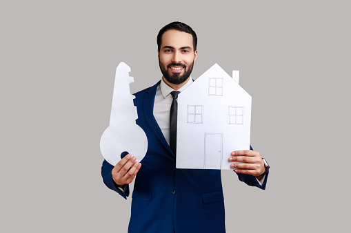 Positive bearded man holding paper house and big key, real estate purchase, rental services, mortgage, wearing official style suit. Indoor studio shot isolated on gray background.