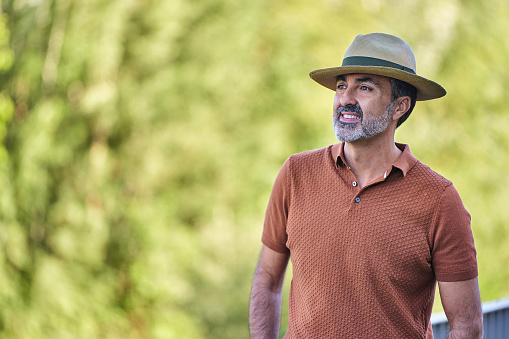 Middle aged man wearing a hat and looking away while standing outdoors against a nature background.