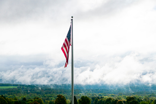 American flag showing it's colors in front of a Cloudy Sky and hills and forest