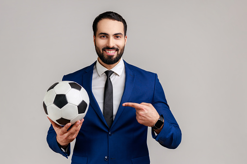 Handsome bearded businessman pointing finger at soccer ball on his hand with smiling positive expression, wearing official style suit. Indoor studio shot isolated on gray background.
