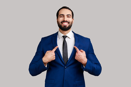 Portrait of bearded man with toothy smile on face pointing fingers on himself, confident positive businessman, wearing official style suit. Indoor studio shot isolated on gray background.