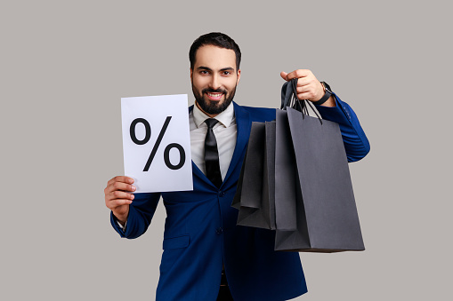 Positive bearded businessman holding shopping bags in hands and paper with percent mark, big discounts, wearing official style suit. Indoor studio shot isolated on gray background.