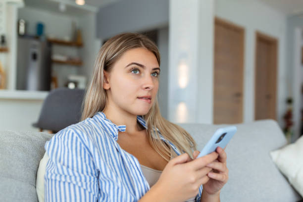 istock Image of nice young serious woman using mobile phone and thinking while sitting on sofa at living room 1428595533