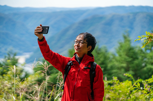 Mid adult man taking a selfie with a smart phone on top of a mountain while hiking in rural Japan