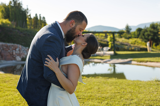 Romantic just married young couple kissing in nature by pond on sunny day