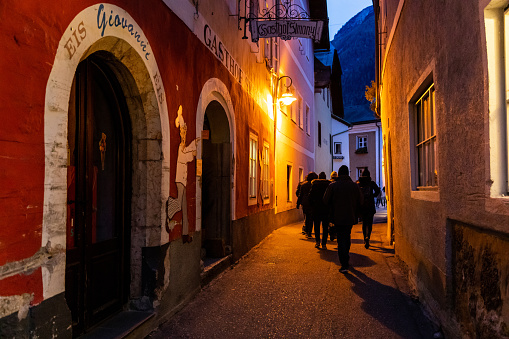 Hallstatt, Austria - November 1, 2019: Tourists walking through small alley in the old town after sunset, most shops and restaurants closed. Small orange street light turned on before night.