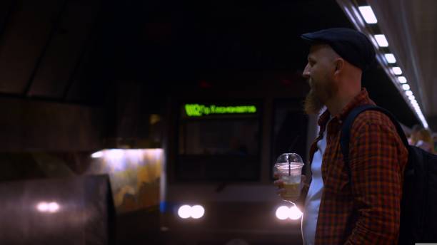 A bearded man in a cap stands on a subway platform and drinks iced coffee while waiting for a subway train. A typical day on the subway, a man goes to work every day early in the morning.