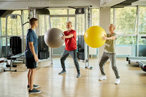 Senior couple exercising with personal trainer using fitness ball at the gym