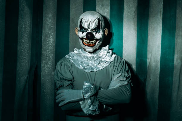 closeup of a creepy bald evil clown, standing in front of a dirty and old circus tent, wearing a gray costume with a white ruff, and starring at the observer with a frightening smile