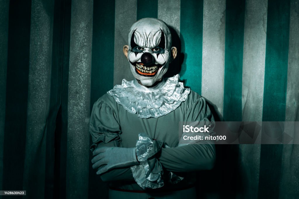 evil clown standing in front of the circus tent closeup of a creepy bald evil clown, standing in front of a dirty and old circus tent, wearing a gray costume with a white ruff, and starring at the observer with a frightening smile Clown Stock Photo