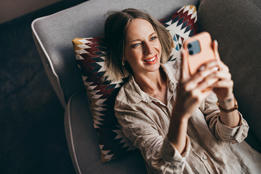 A from above view of a smiling Caucasian entrepreneur holding her smartphone and texting somebody while sitting on her cozy sofa.