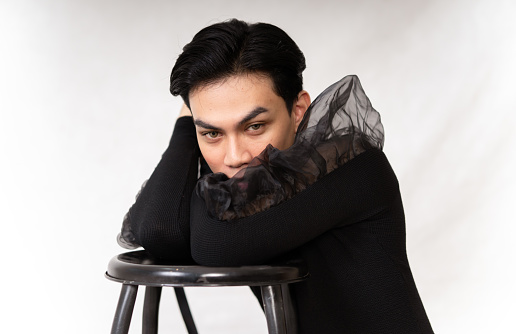 Pretty Asian man putting his hand and head on the chair while posing in a black dress inside the studio