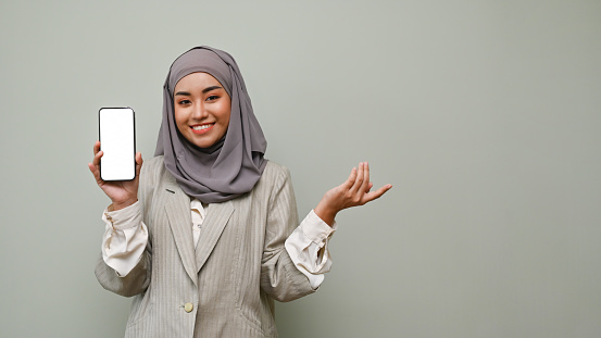 Attractive and beautiful young Asian Muslim woman wearing hijab showing a smartphone white screen mockup, standing isolated over gray studio backdrop.