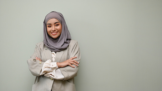 Beautiful and confident young Asian Muslim woman or businesswoman wearing hijab stands with her arms crossed against a grey studio background. copy space on grey background to show your text
