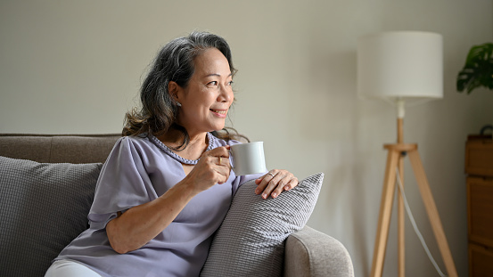 Happy and calm Asian 60s aged-woman sipping her morning coffee while relaxing in her living room, looking out the window with happy expression.