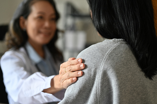 An aged Asian female doctor touching shoulder to comfort and support her patient. A young Asian female patient is being reassured by her doctor. close-up image