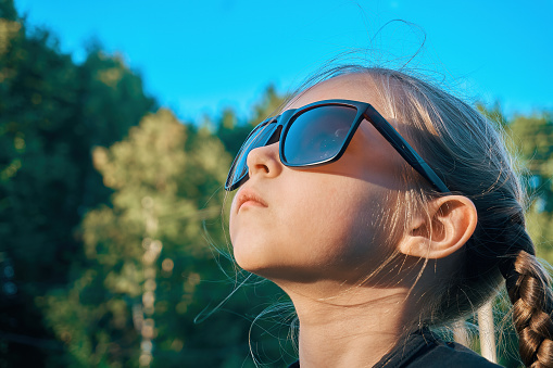 A girl child in sunglasses looks at the top in the direction of the sun. Selective soft selective focus.