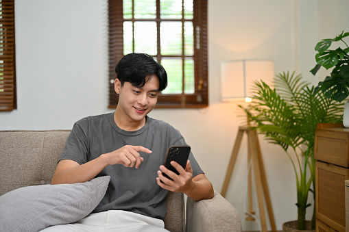 Handsome young Asian man using smartphone while relaxing sitting on the couch at home. enjoying new application, scrolling on online shopping website, chatting with friends.