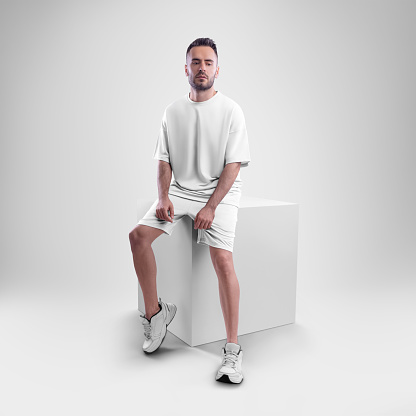 Mockup of white men's oversized t-shirt with shorts on a man sitting on a cube. Clothing template for presentations of design, print, pattern.