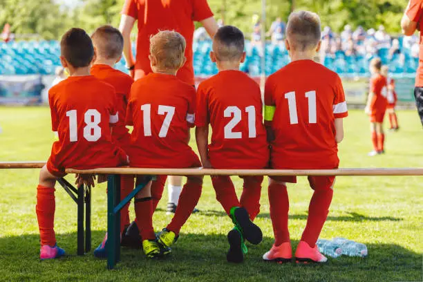 Photo of Football players in youth team sitting on wooden bench. Kids in red shirts in school sports team. Boys watching tournament game at soccer stadium