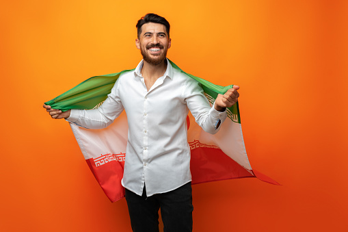 Portrait of a young casual man holding flag of Iran against orange background in studio