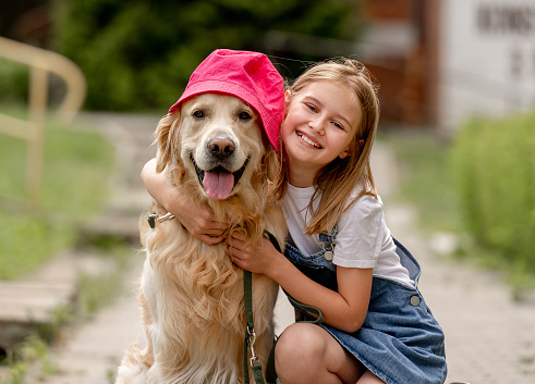 Preteen girl wearing hat hugging golden retriever dog sitting outdoors in summertime. Pretty kid petting fluffy doggy pet in city