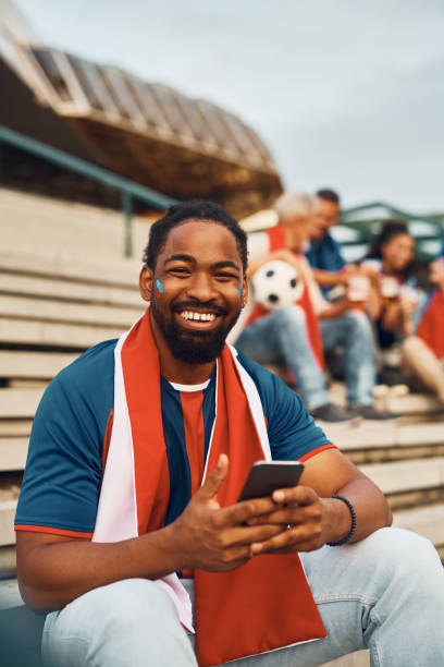 istock Happy black soccer fan using smart phone on a stadium and looking at camera. 1428669527
