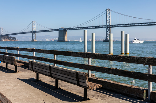 Bench near fence on edge of waterfront beach with view of San Francisco - Oakland Bay Bridge of sunny day in California, USA.