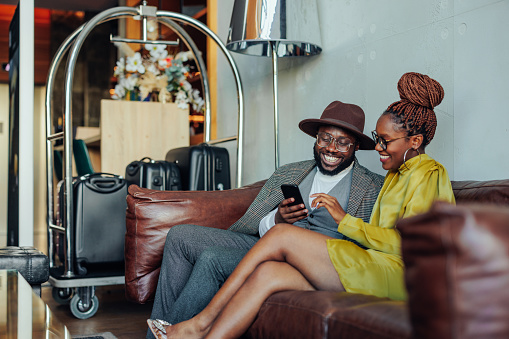 A wealthy couple is lounging in the hotel as they're waiting to be taken to their luxurious suit in the hotel. They are sitting on a sofa with their luggage on the cart and are using a smartphone.