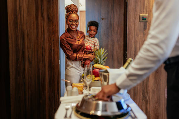 Wealthy hotel guest receiving room service. A wealthy African American hotel guest is with her son at the apartment door, she is being catered with lavish room service by the waiter. room service stock pictures, royalty-free photos & images