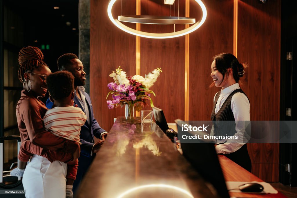 Busy front desk at hotel. A hotel clerk is welcoming guests that are checking in. It is a wealthy African American couple with their baby boy. The smiling clerk tends to them. Hotel Stock Photo
