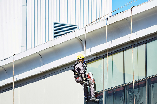 Cape Town, South Africa - 27 September 2022: Maintenance worker hanging from a rope as he abseils on the exterior of a commercial building, wearing a yellow hard hat, overalls, gloves and boots, in Claremont, Cape Town, South Africa.