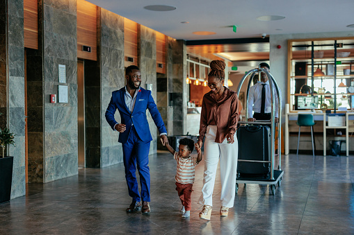 A hotel clerk is welcoming guests that are checking in. It is a wealthy African American couple with their baby boy. The smiling clerk tends to them.