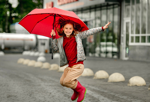 Preteen girl wearing roober boots and holding umbrella jumping in rainy autumn day and smiling. Pretty child in gumboots having fun at wet street