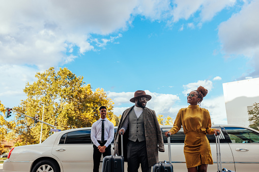 A stylish African American couple arrive at the airport. They are carrying their luggage, as their chauffeur is standing behind next to their limousine.