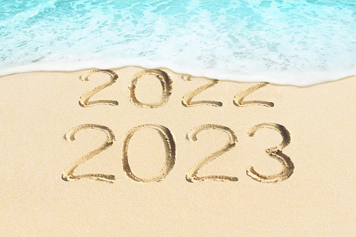 Years numbers 2022 and 2023 handwritten in sand. Soft ocean wave splashing on sandy beach background.