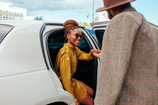 A happy African American couple is on their luxurious vacation. They are entering a limousine waiting for them. The husband is helping his wife entering the car.