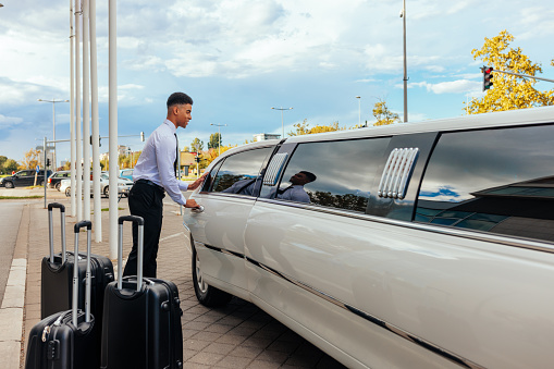 A young African American chauffeur is closing the door of the limousine after his clients entered the vehicle. There are some suitcases standing next to the car.