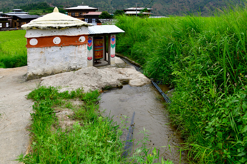 Youwakha, Lobesa valley, Barp Gewog, Punakha District, Bhutan: water powered prayer wheel - small temple in the middle of the rice fields - the water flow from the nearby brook provides hydraulic energy to power the prayer wheel. A prayer wheel, also known as a mani-mill or mani-wheel, is a cylinder containing prayers or mantras printed on or decorated on the outside of a roll of paper. Prayer wheels commemorate the turning of the wheel of teaching (dharmachakra) by Buddha himself and are intended to open up opportunities for those who are illiterate to acquire positive karma .