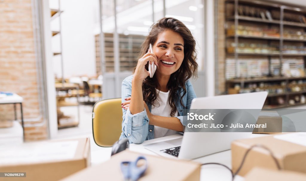 Happy young businesswoman speaking on the phone in a warehouse Happy young businesswoman speaking on the phone while working in a warehouse. Online store owner making plans for product shipping. Creative female entrepreneur running an e-commerce small business. Using Phone Stock Photo