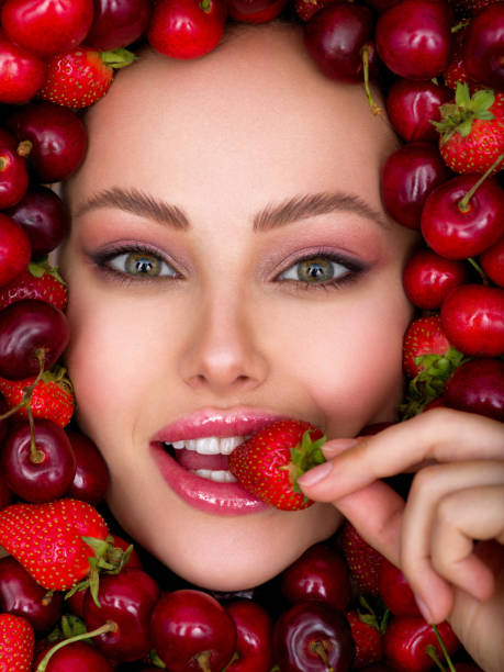 Young girl with bright makeup and a berry background.  Attractive woman tasting cherry and strawberry. Young girl with bright makeup and a berry background. Beautiful caucasian woman biting a berry. Attractive woman tasting strawberry. Beautiful woman with fashion makeup and a vivid background. tasting cherry eating human face stock pictures, royalty-free photos & images