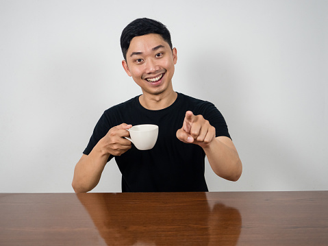 Positive man smile and hold coffee cup sit at table point finger at you