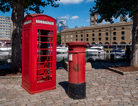 A traditional red British telephone box and postbox standing on a cobblestone area in front of a collection of boats moored in St Katharine Docks near Tower Bridge in London. The buildings to the right is an old warehouse, now converted into apartments.